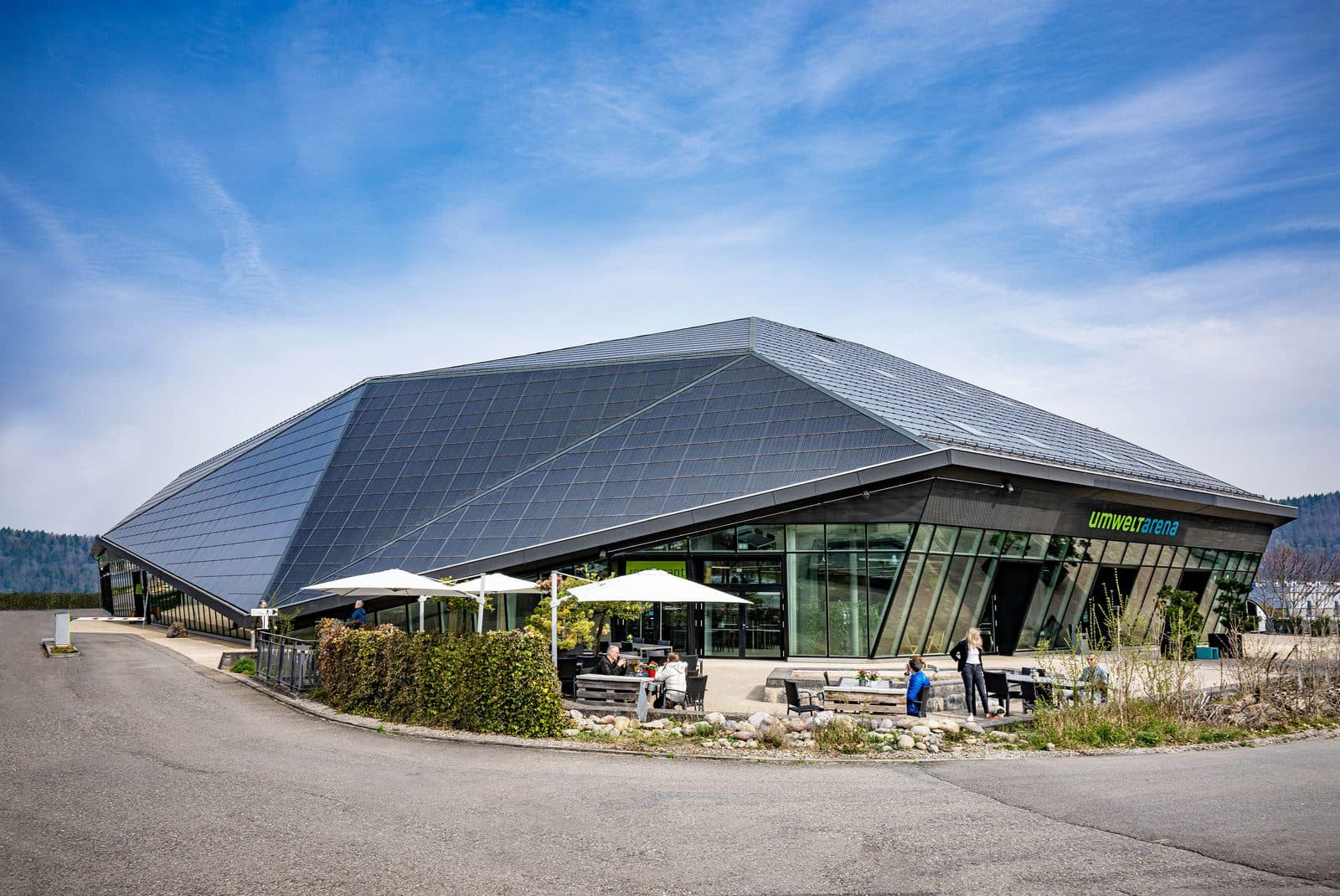 Die Umwelt Arena Schweiz in Spreitenbach: ein Gebäude mit grossem, schrägem Solardach, davor eine Terrasse mit Sonnenschirmen.